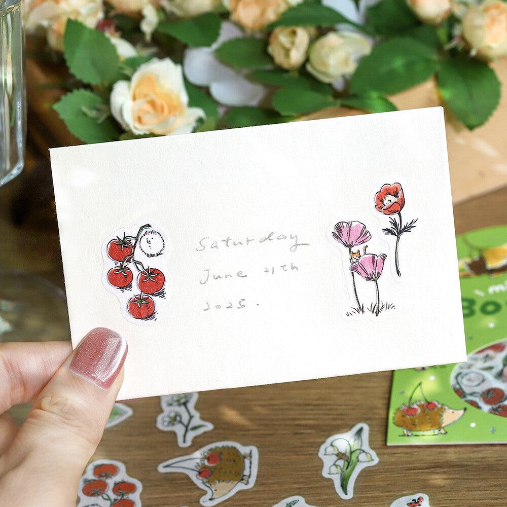 Hand holding a card with illustrations of tomatoes and flowers on a wooden surface with more cards and flowers in the background.
