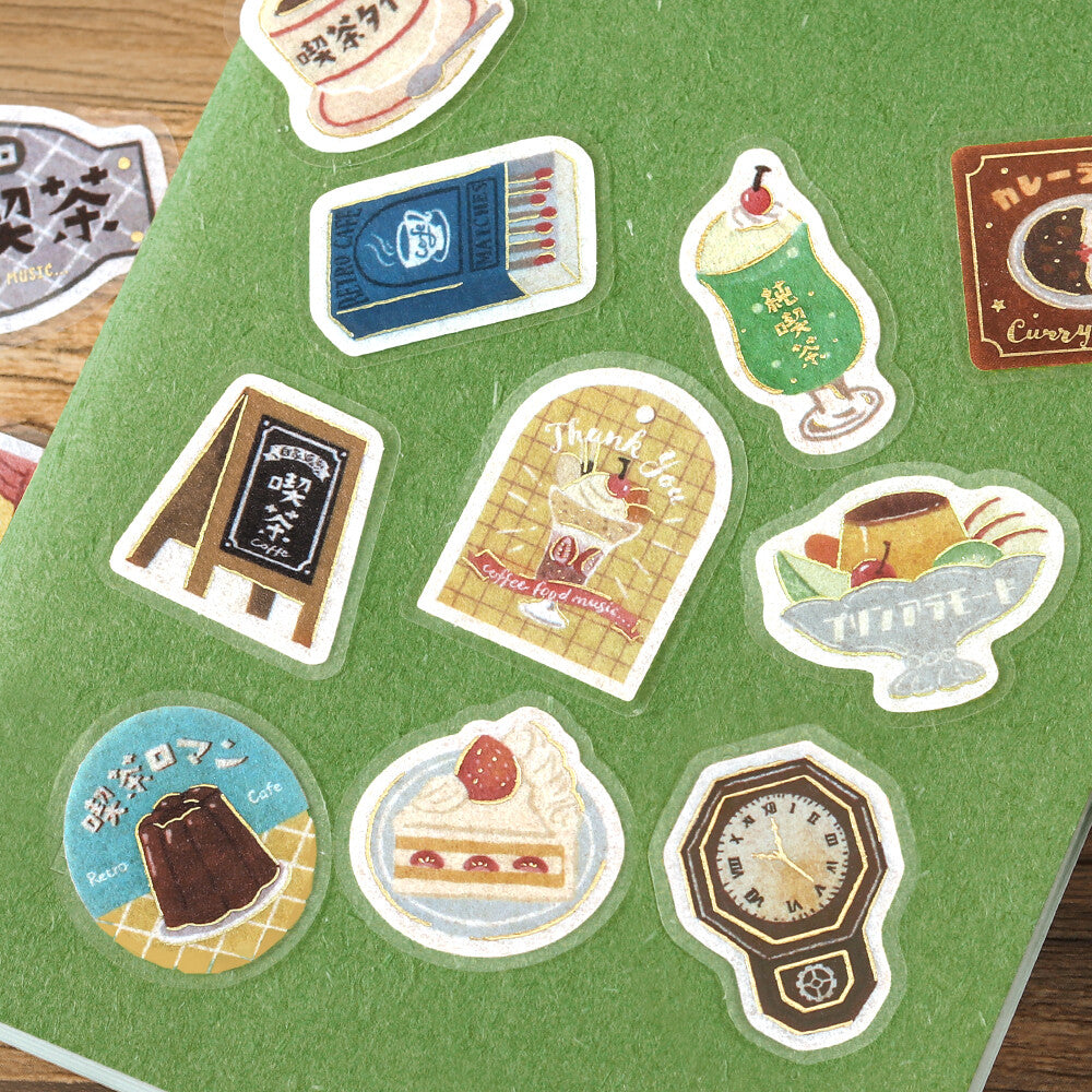 A collection of cute, illustrated die-cut stickers displayed on a green felt sheet over a wooden surface. The stickers feature various whimsical designs including a green milk tea drink, a strawberry cake slice, a chocolate dessert, a retro clock, a sign, and other food and object illustrations.