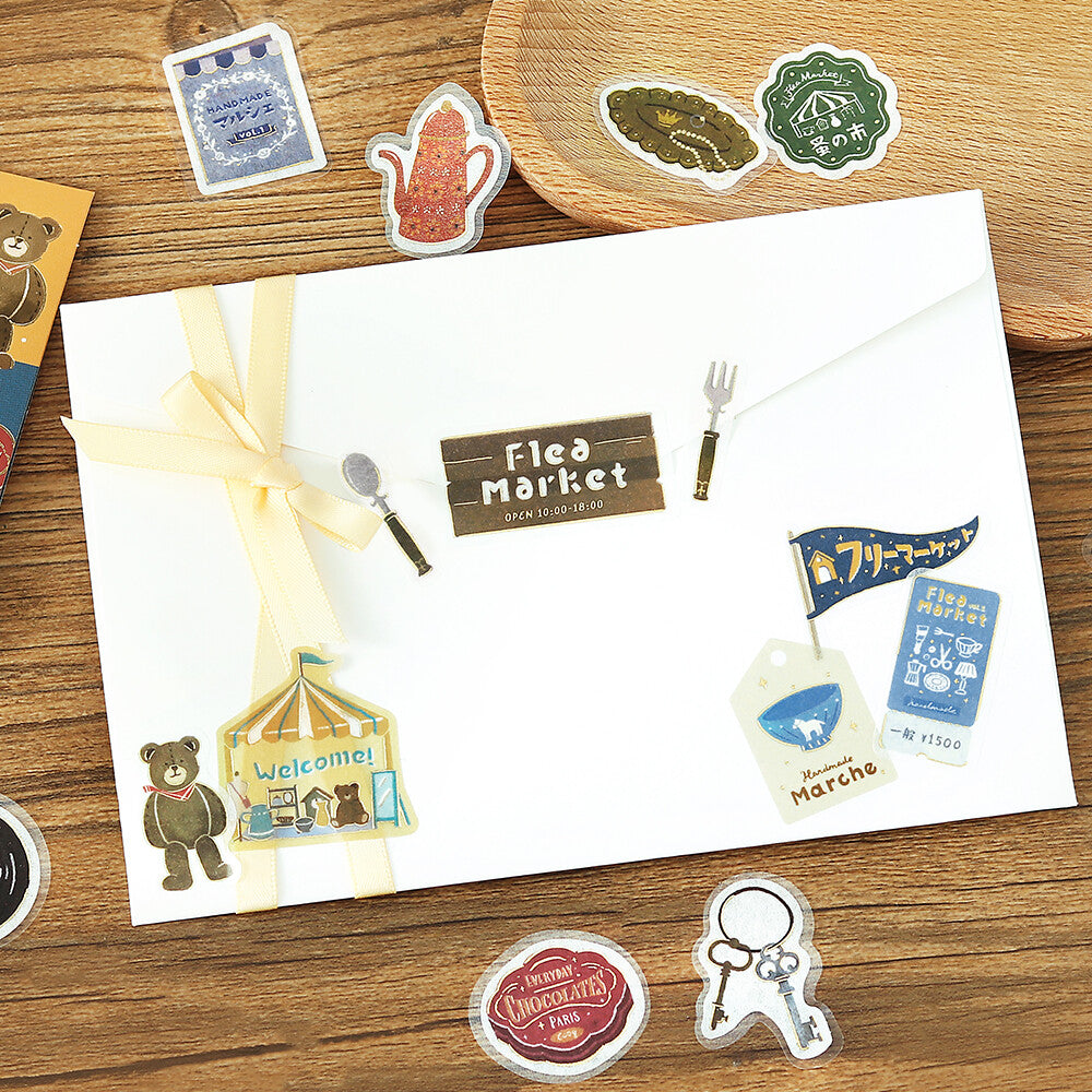 A white envelope decorated with a pale yellow ribbon, small decorative stickers, and a wooden-style 'Flea Market' label, arranged on a wooden tabletop with other assorted stickers around it.