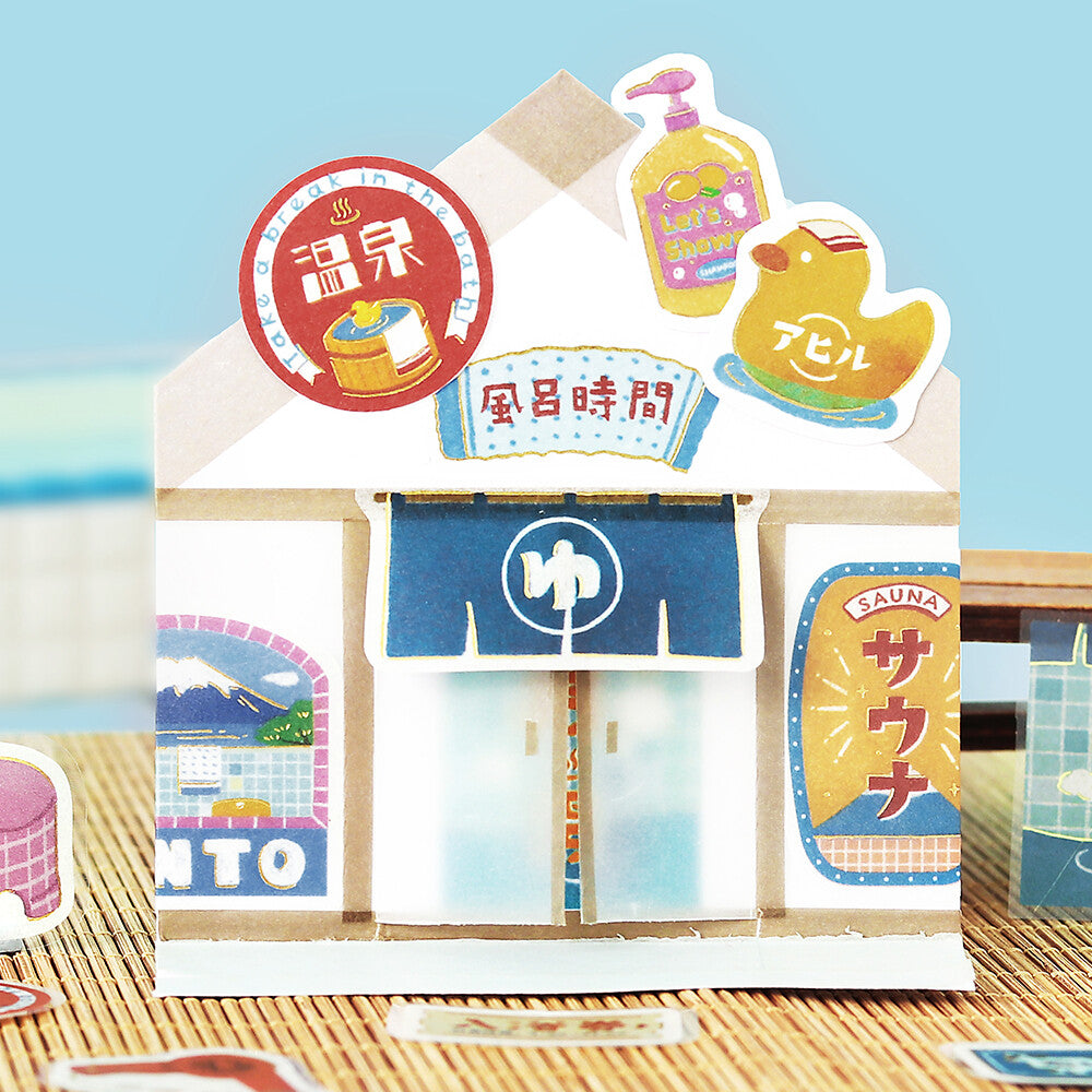 A miniature paper-style hot spring and sauna shop display with colorful illustrated signs, a blue awning, and small decorative props on a tabletop against a soft blue background.