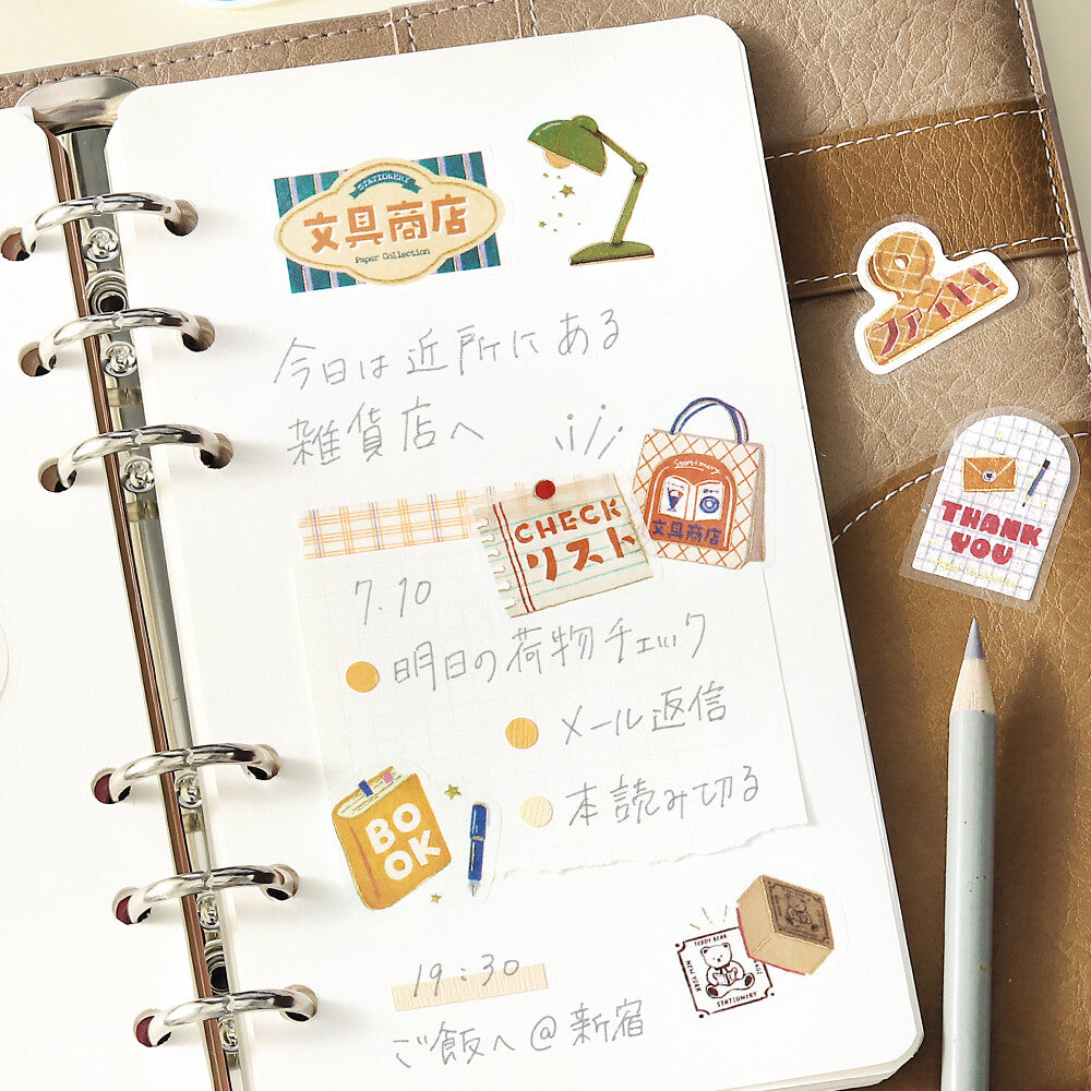 A white spiral-bound planner or notebook page decorated with cute Japanese stationery stickers, handwritten notes, and a pencil on a brown textured leather surface.