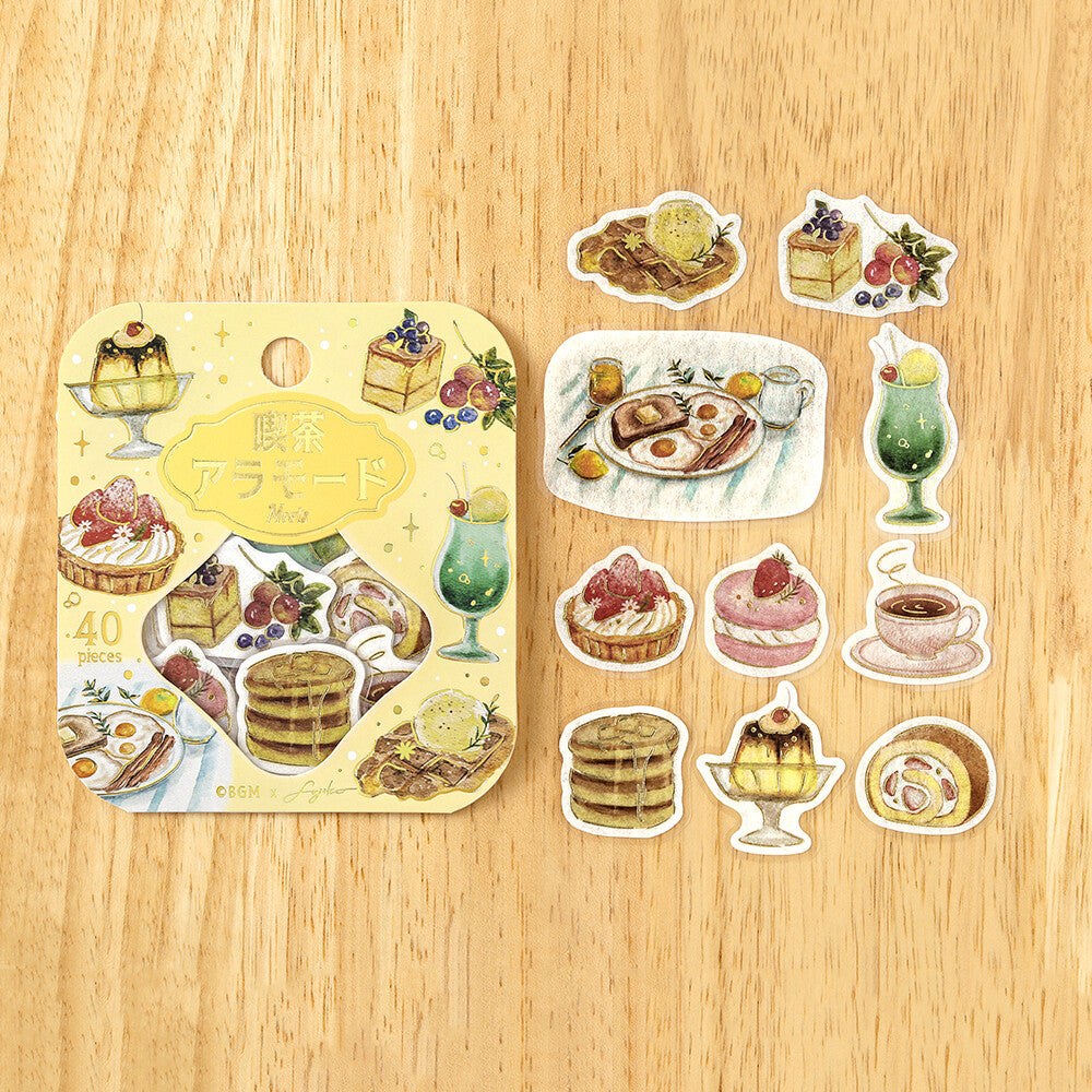 A sheet of illustrated food stickers displayed on a light wood surface, alongside several individual sticker designs showing desserts, drinks, and breakfast scenes.