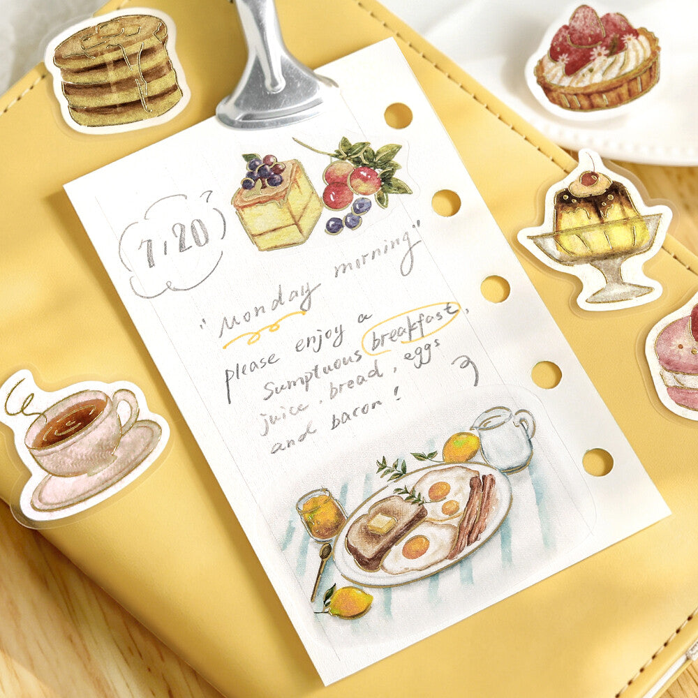 A sheet of die-cut planner stickers featuring watercolor breakfast illustrations and a handwritten Monday morning quote, arranged on a yellow notebook.