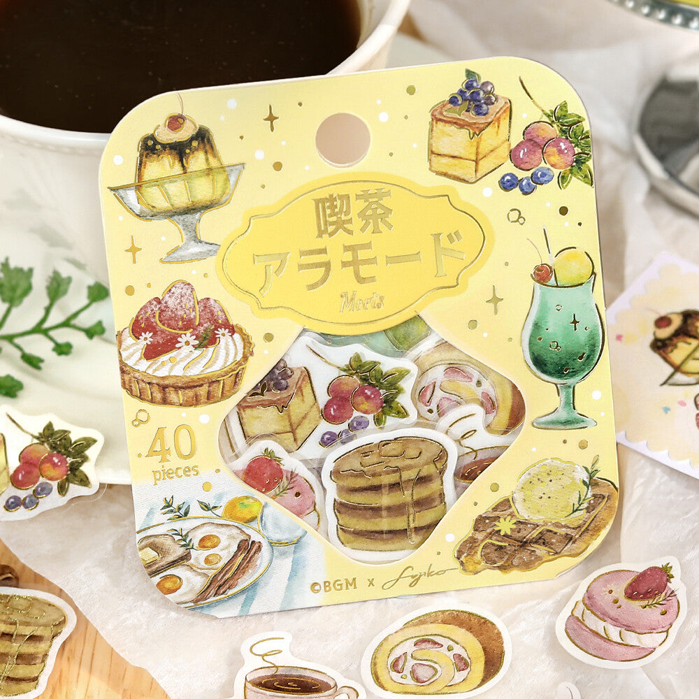 A packaged set of food-themed stickers or decals on a yellow backing card, surrounded by loose sample stickers. The card shows illustrated desserts, pancakes, fruit, and drinks with Japanese text and the visible brand mark 'OGM x Sugar'.
