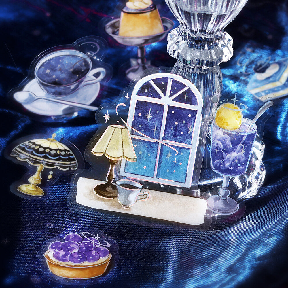 A collection of illustrated acrylic charms or stickers depicting a cozy night scene, including a window with a starry blue sky, a glowing lamp, a teacup, a glass of purple iced tea with a yellow lemon, and a purple berry tart, arranged on a dark blue satin fabric background.