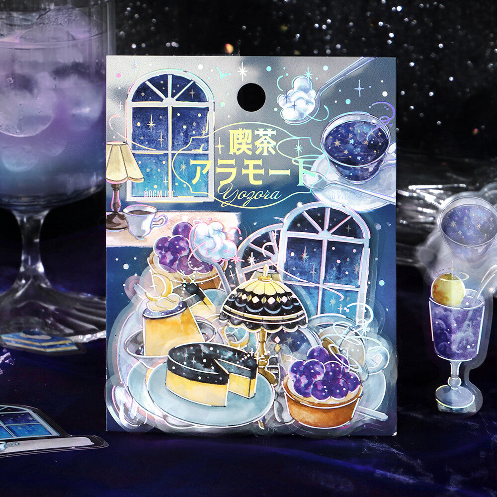 A decorative printed card or packaging insert featuring a whimsical illustrated winter tea scene with windows, cups, and desserts, placed on a dark blue surface with blurred drink glasses in the background.