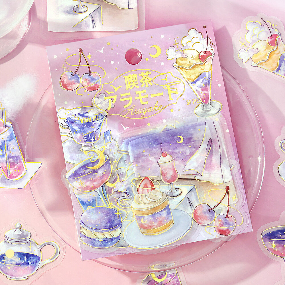 A pastel pink illustrated sticker sheet or sticker pack featuring desserts, teacups, cherries, and celestial motifs, displayed on a clear round plate with matching stickers around it.