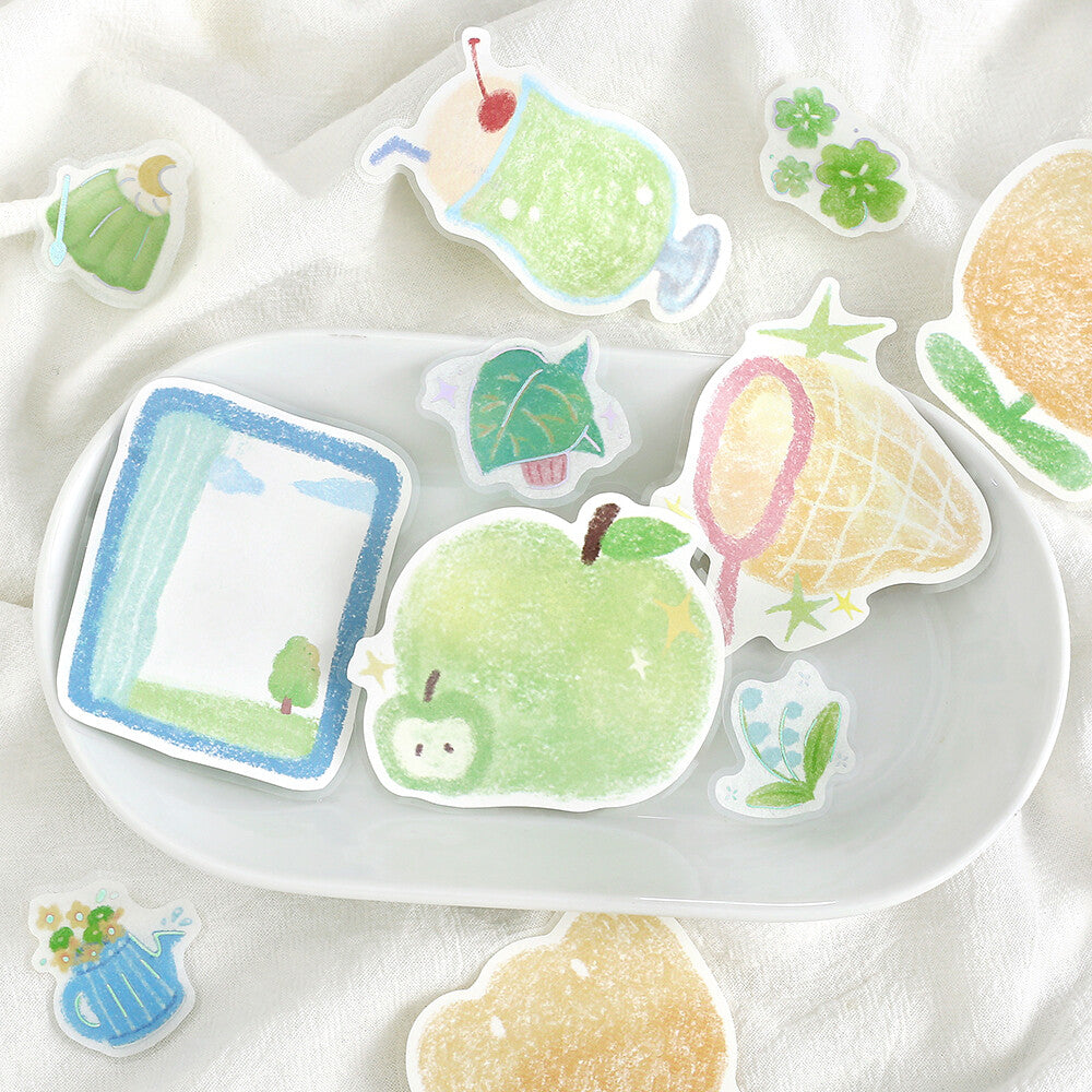 A collection of cute, die-cut sticker illustrations arranged on a white ceramic plate over a soft white fabric background. The stickers feature whimsical drawings including a green apple, a pink and yellow ice cream cone, a blue window, a green leaf, and a blue watering can.