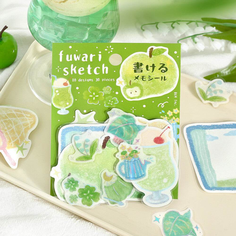 A package of fuwari sketch memo stickers displayed on a beige tray, surrounded by loose stickers featuring green-themed illustrations like drinks, flowers, and leaves.