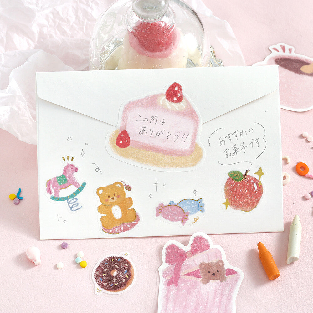 A white envelope decorated with cute pastel stickers and Japanese text, arranged on a pink surface with small candy-like props and a glass dome in the background.