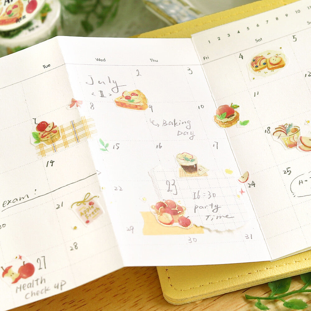 A close-up of an open planner or notebook showing decorated weekly calendar pages with watercolor illustrations of pastries, fruit, and small stickers, placed on a yellow notebook over a wooden surface.