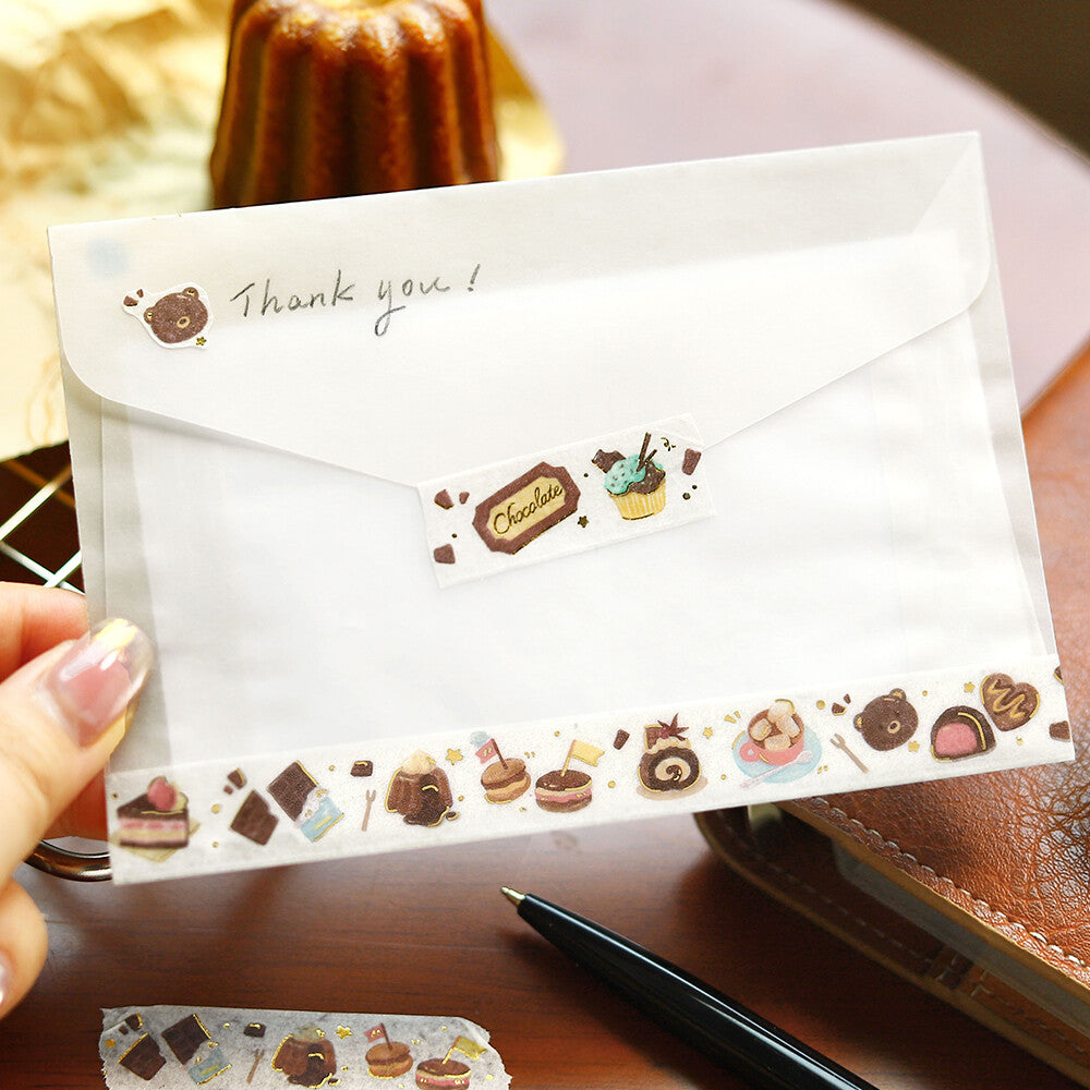 White envelope with chocolate-themed stickers and 'Thank you!' text, held by a hand on a wooden surface.