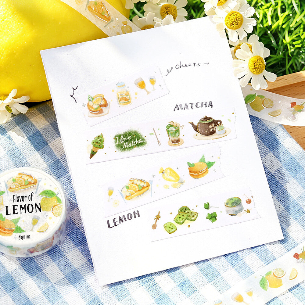 A sheet of decorative washi tape with lemon and matcha-themed illustrations is displayed on a blue-and-white checkered cloth with daisies, a lemon, and a roll of 'Flavor of Lemon' washi tape nearby.