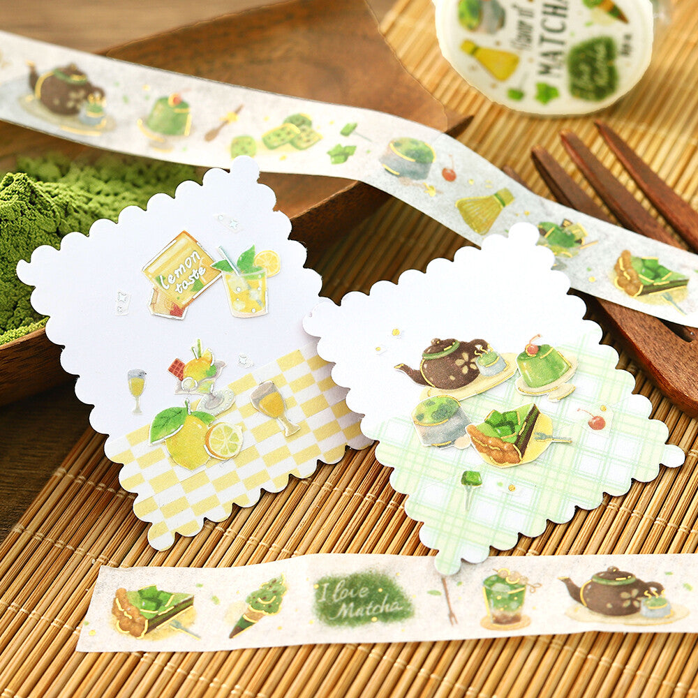 Three scalloped white decorative stickers or labels with green tea-themed illustrations, displayed on a bamboo mat with a matching roll of tea-patterned tape in the background.