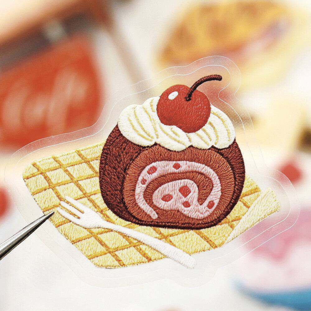 A close-up of an embroidered patch or applique depicting a dessert roll cake with whipped cream, a cherry on top, and a small white fork, shown against a softly blurred background.