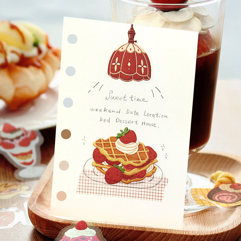 A vertical cream-colored greeting card with a red and gold illustration of a dessert lamp and a waffle with strawberries, reading 'Sweet time weekend Date Location Red Desert House.' The card is photographed standing upright in a styled tabletop setting with a drink, pastries, and small decorative stickers around it.