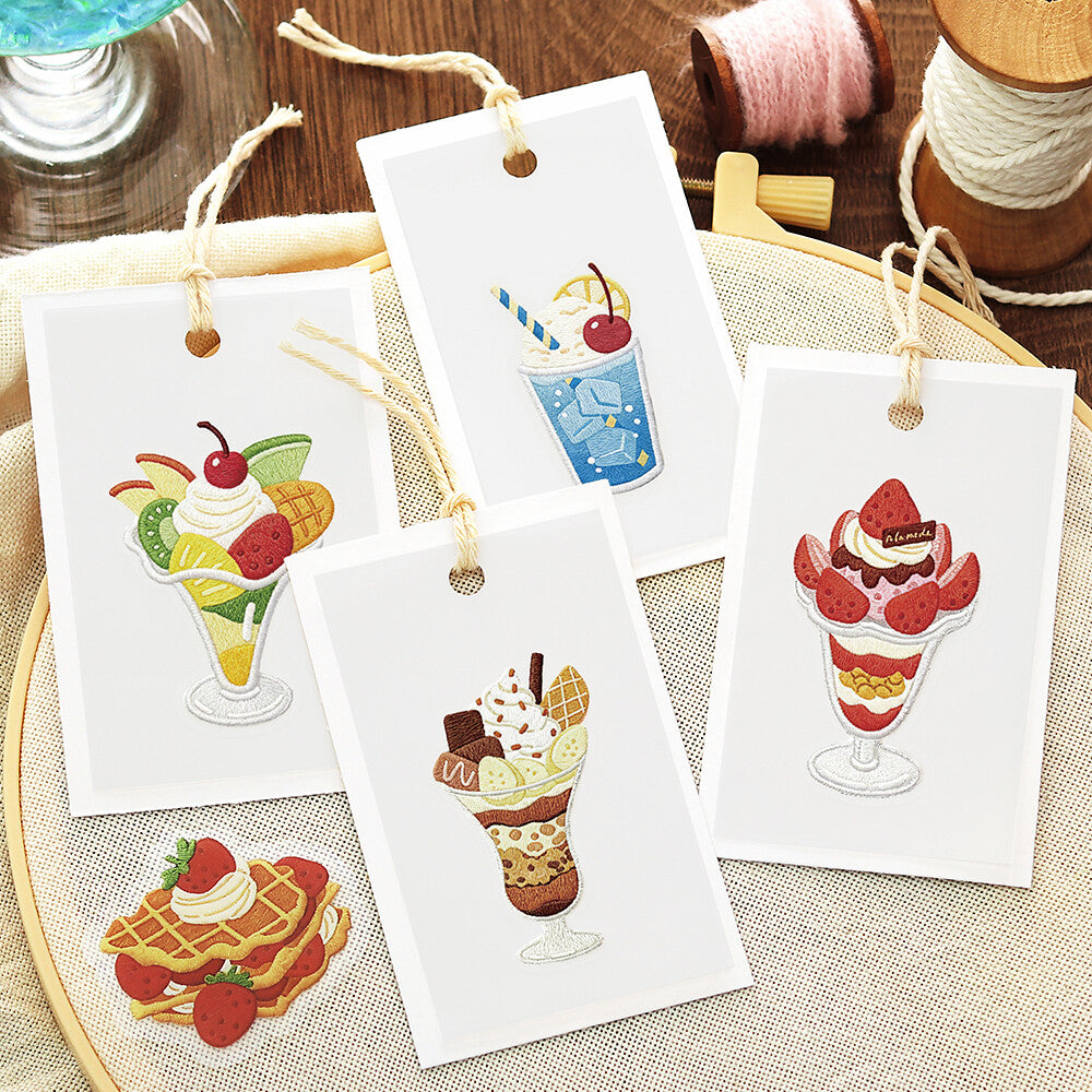 Four white rectangular tags with illustrated dessert drawings and hanging twine loops, arranged on a woven fabric surface with spools of thread in the background.