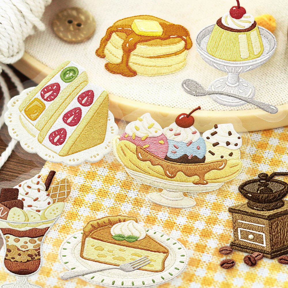 A collection of embroidered dessert and pastry motifs arranged on a yellow-and-white checkered fabric surface, including pancakes with syrup, a pudding on a pedestal, layered cakes, ice cream sundaes, a pie slice, and small coffee-themed accessories.