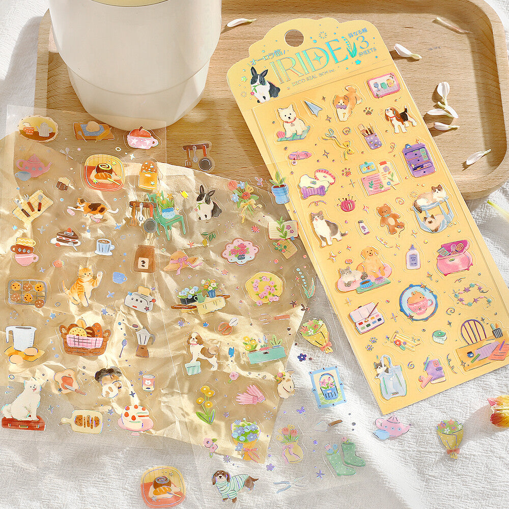 Two sheets of cute, colorful cartoon stickers featuring animals, food, and everyday objects, displayed on a light fabric surface next to a wooden tray and a white mug.