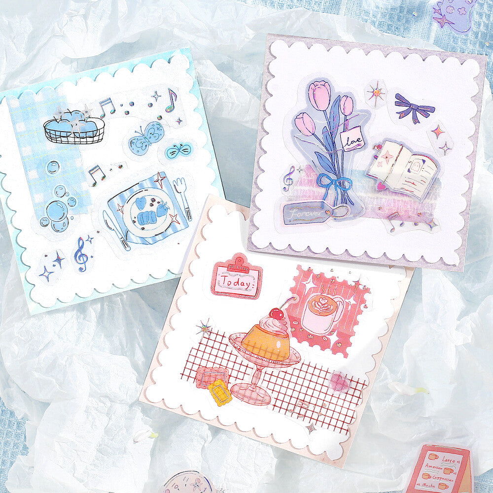 Three decorative square cards with scalloped edges are arranged on a crinkled white fabric background. The cards feature pastel illustrations: one blue music-themed design, one pink tulip and book design, and one orange dessert design.