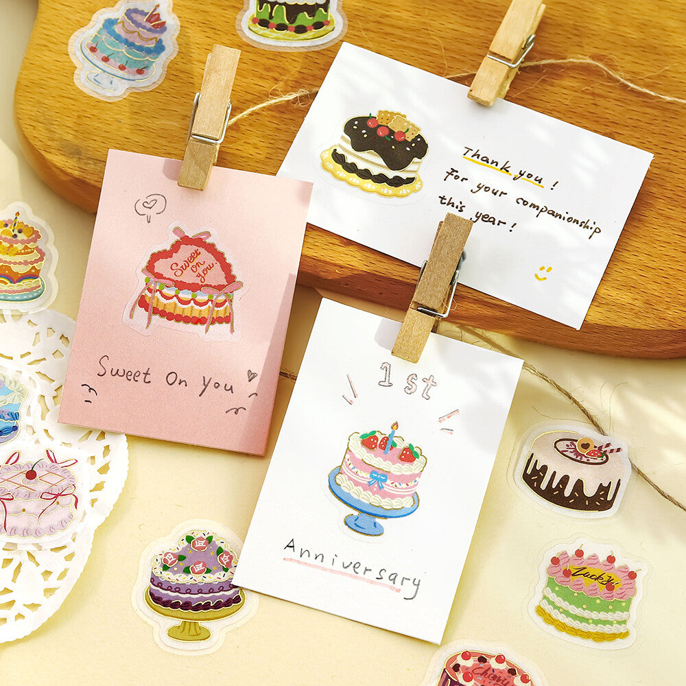 A styled flat-lay of cute, colorful cake-themed stickers on a wooden board and cream fabric background, with two cards held by wooden clothespins.