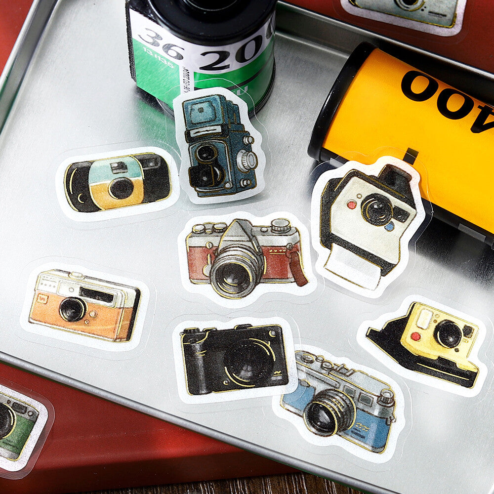 Collection of camera-shaped stickers on a metallic surface with film and camera equipment in the background.