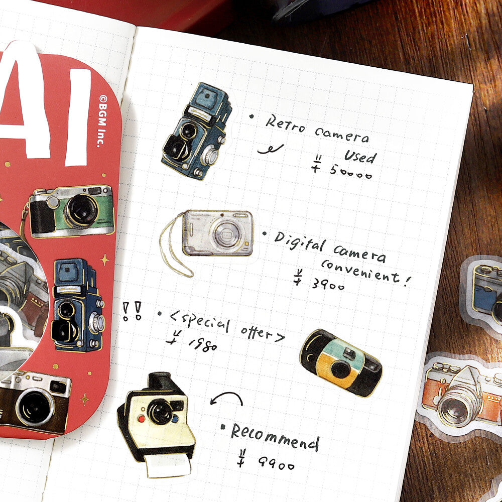 A styled flat-lay of a white graph paper notebook page featuring retro camera illustrations and handwritten notes in Japanese, alongside a red decorative card and small camera sticker decals on a wooden surface.