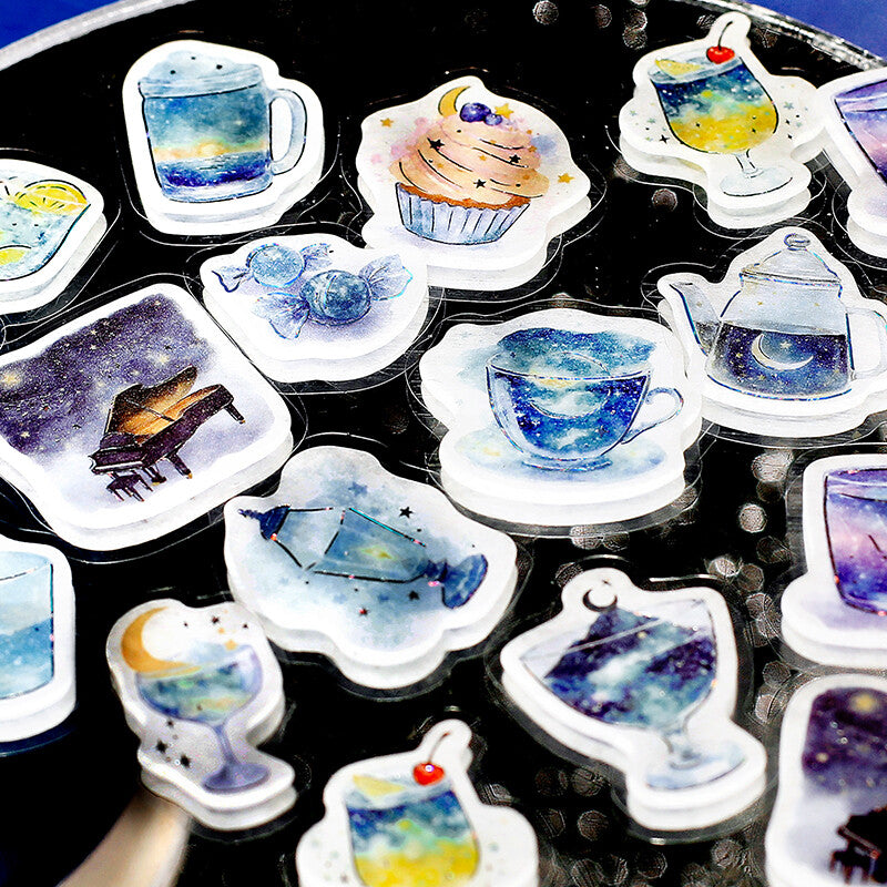 A collection of die-cut acrylic stickers featuring watercolor illustrations of celestial-themed designs, including a galaxy cupcake, a galaxy cocktail, a galaxy teapot, and a galaxy piano, arranged on a dark reflective surface.