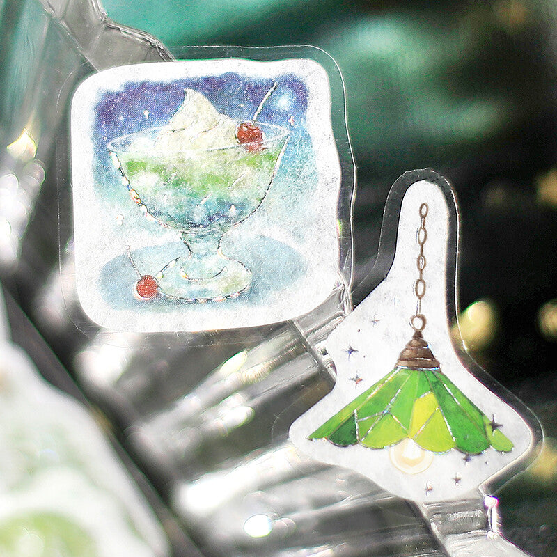 Two clear acrylic hanging ornaments with printed illustrations, one showing a dessert glass with cherries and the other a green hanging lamp, photographed against a blurred dark green background.