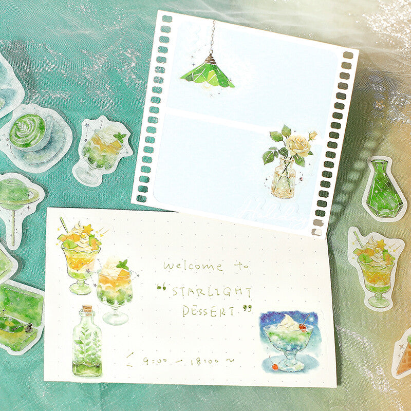 A styled flat-lay of decorative stationery featuring watercolor illustrations of desserts, drinks, and small objects. Two white paper sheets with perforated edges are arranged on a textured teal surface, surrounded by matching die-cut stickers.