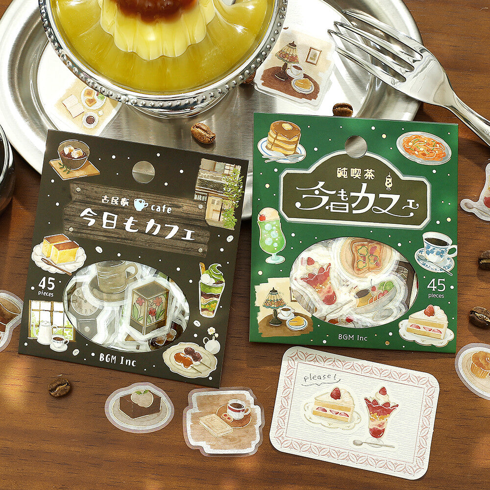 Two illustrated sticker sheets and several loose die-cut stickers are arranged on a wooden table with a dessert cup, fork, and coffee cup props. The packaging features cafe-themed illustrations and Japanese text, with visible brand text 'BGM Inc' on the boxes.