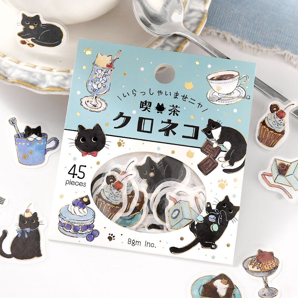 Pack of decorative stickers with black cat illustrations on a table with tea cups and a spoon.