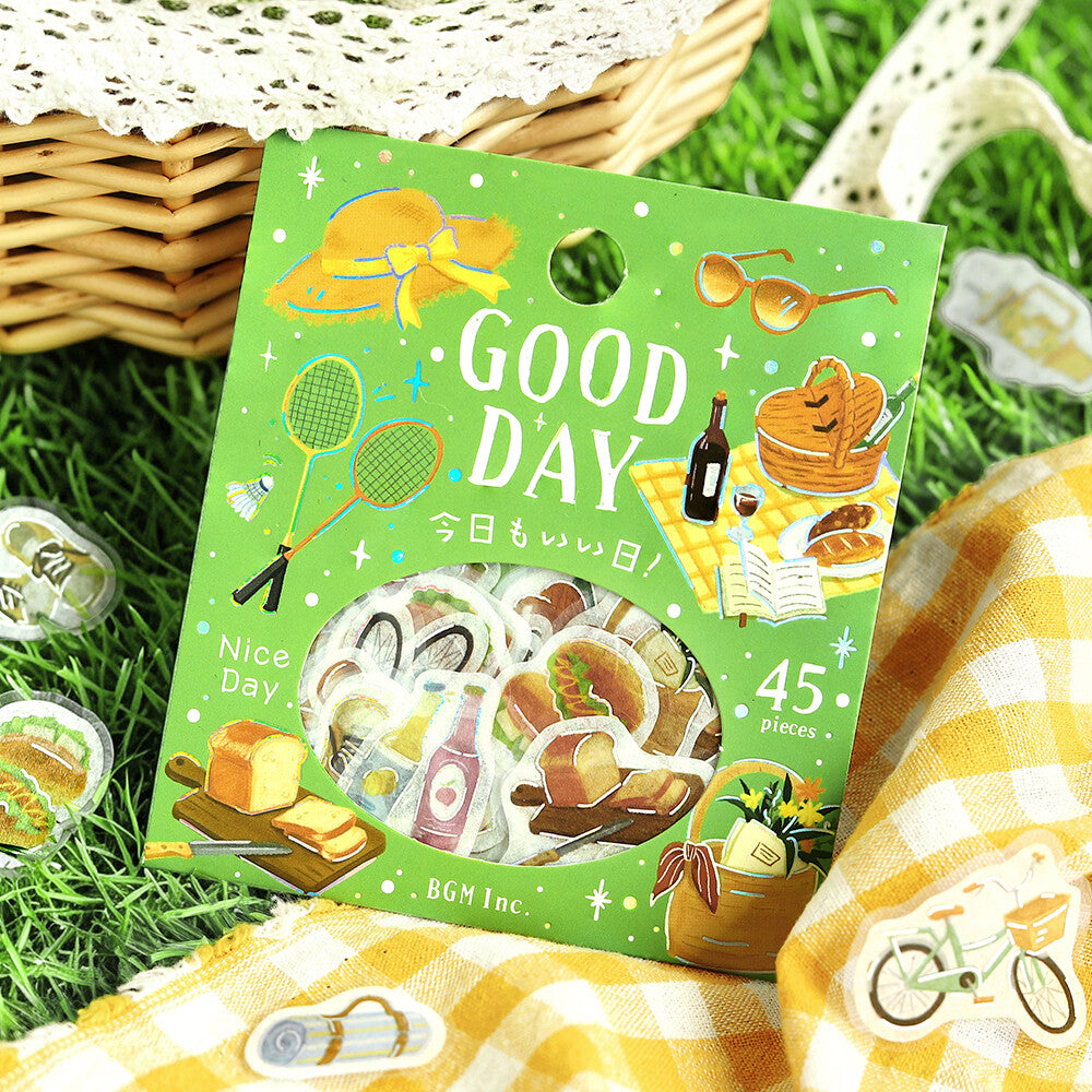 A green sticker pack box labeled 'GOOD DAY' with picnic-themed illustrations and a circular window showing assorted stickers, placed on grass with a yellow-and-white check blanket, a woven basket, and a lace doily around it.