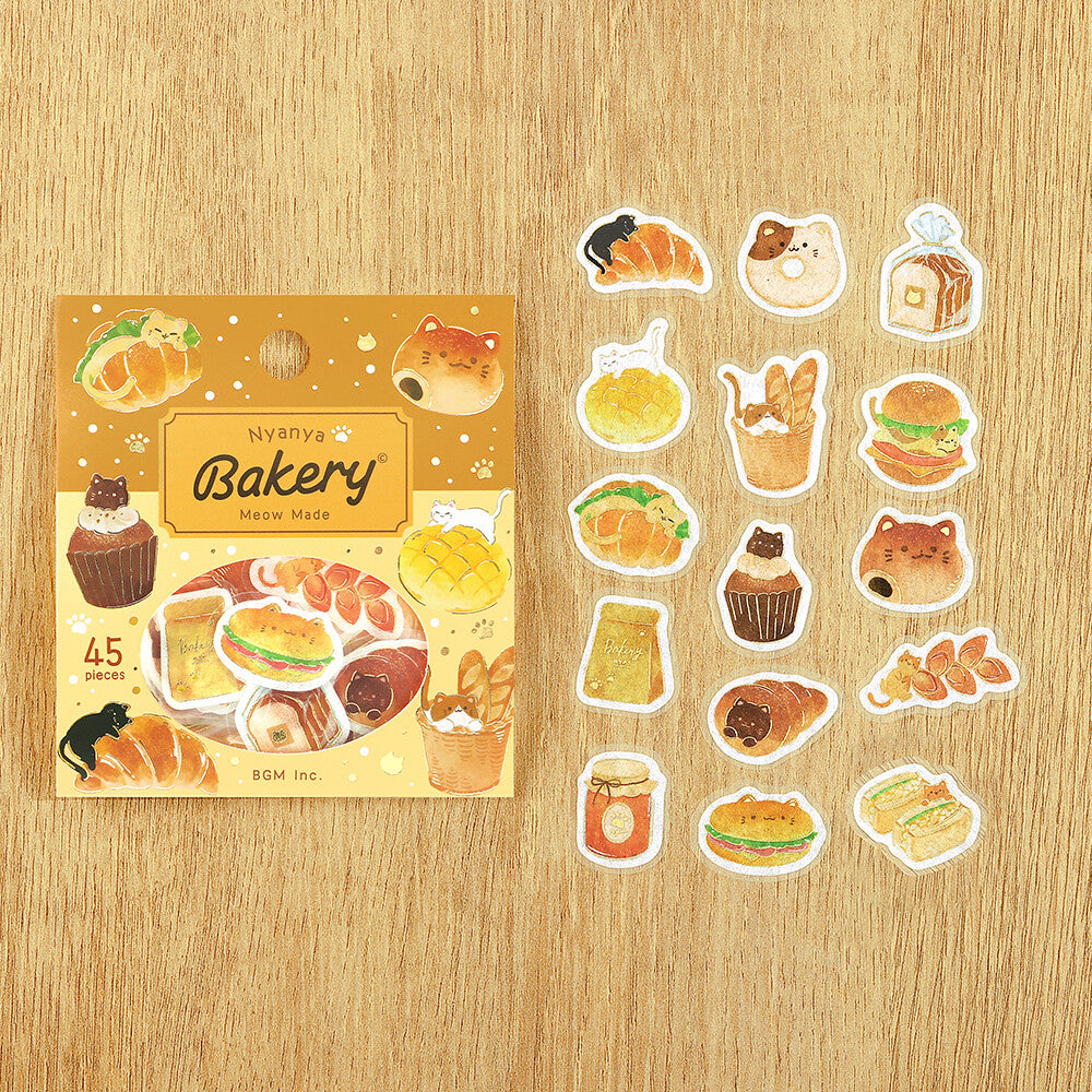 A sheet of 12 bakery-themed die-cut stickers displayed next to their original packaging on a light wood grain background. The packaging is yellow with illustrations of pastries and the brand name 'banya' is visible.
