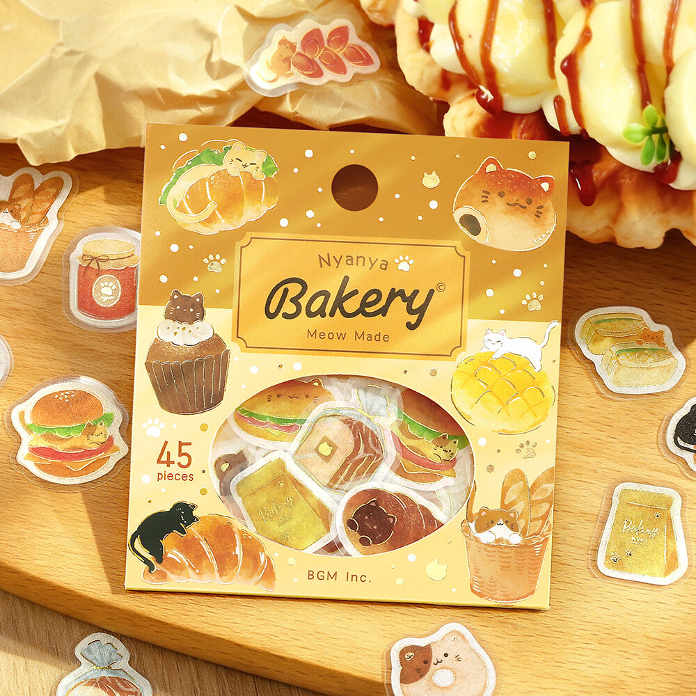 Nyanya Bakery Meow Made sticker pack on a wooden tabletop with waffles and food props in the background. The yellow package contains 45 assorted bakery-themed stickers shown through a clear window.