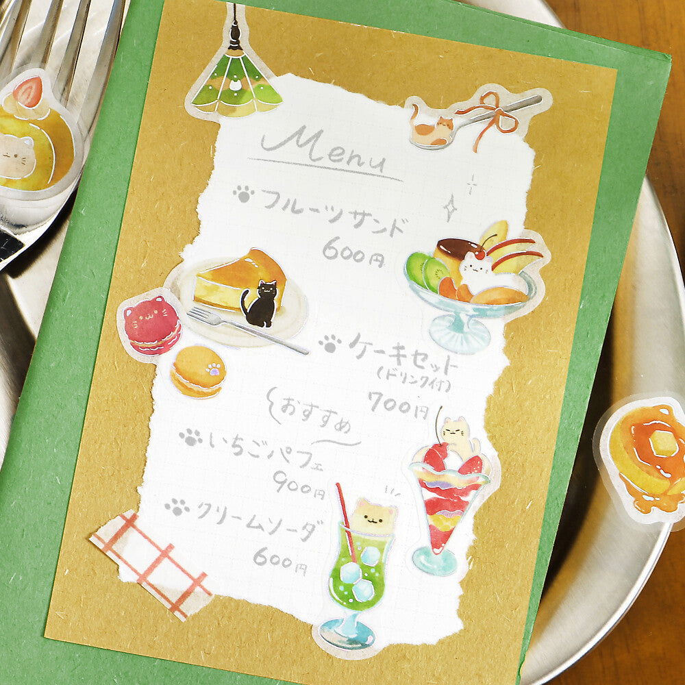 A close-up of a cute illustrated menu card with Japanese text and whimsical dessert illustrations, placed on a green folder over a white plate with utensils nearby.