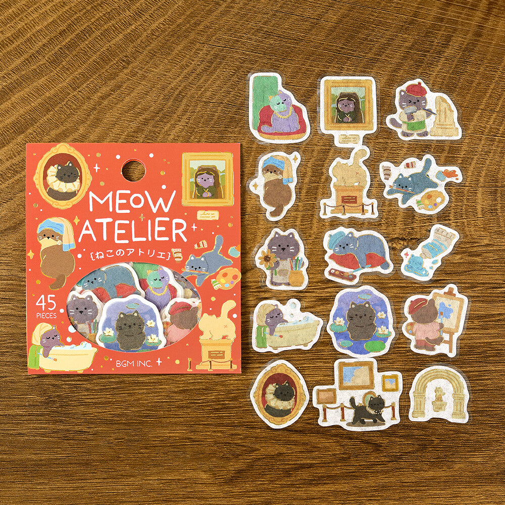 A set of cute cat-themed stickers displayed next to their red packaging card on a wooden tabletop. The packaging is labeled 'MEOW ATELIER' and '45 PIECES', featuring illustrations of cats in cozy home scenes.