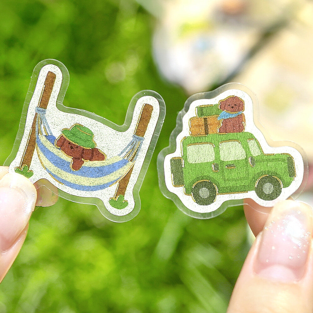 Two clear acrylic stickers held by fingers against a blurred green outdoor background. One sticker shows a brown dog relaxing in a blue and white hammock with a green hat, and the other shows a green car carrying a brown dog with luggage on top.