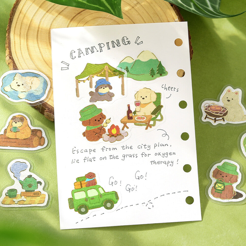 A sheet of cute camping-themed planner stickers displayed on a green textured background with a wood slice prop. The sheet features illustrations of a bear in a green hat setting up a tent, a campfire, a green van, and handwritten text, surrounded by individual die-cut sticker designs.