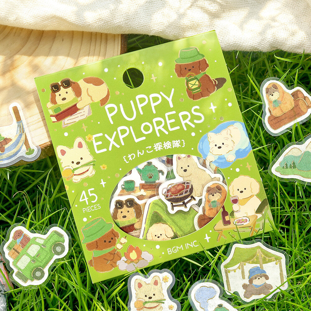 A package of 'Puppy Explorers' sticker stickers by BGM INC, displayed on a green grass background with a beige fabric cloth. The packaging is light green and features cute cartoon puppy illustrations.