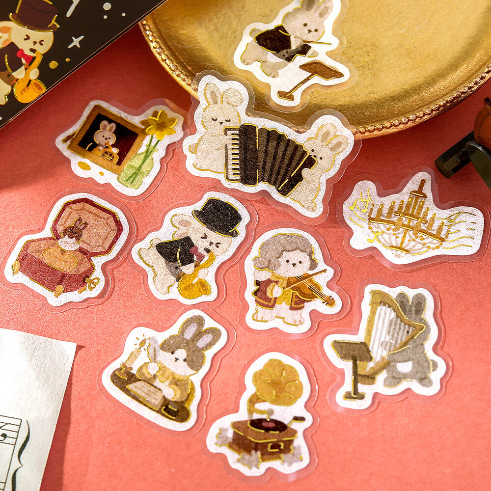 A set of cute embroidered bunny-themed stickers displayed on a coral pink background with a gold plate prop. The stickers feature various bunnies playing musical instruments like an accordion, harp, violin, and gramophone, along with cozy scenes.