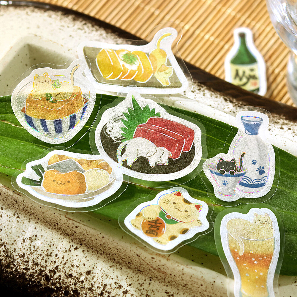 A set of die-cut sticker illustrations featuring cute cartoon cats in Japanese-themed settings, displayed on a green leaf over a speckled ceramic dish.