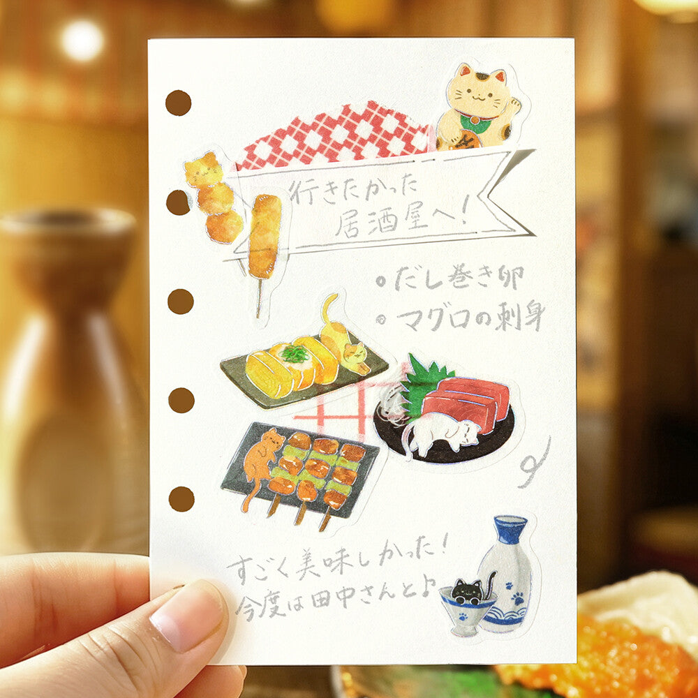 A hand holds a white sticker sheet featuring cute Japanese food illustrations, including sushi, yakitori, sashimi, and a sake bottle, against a warm, blurred background.
