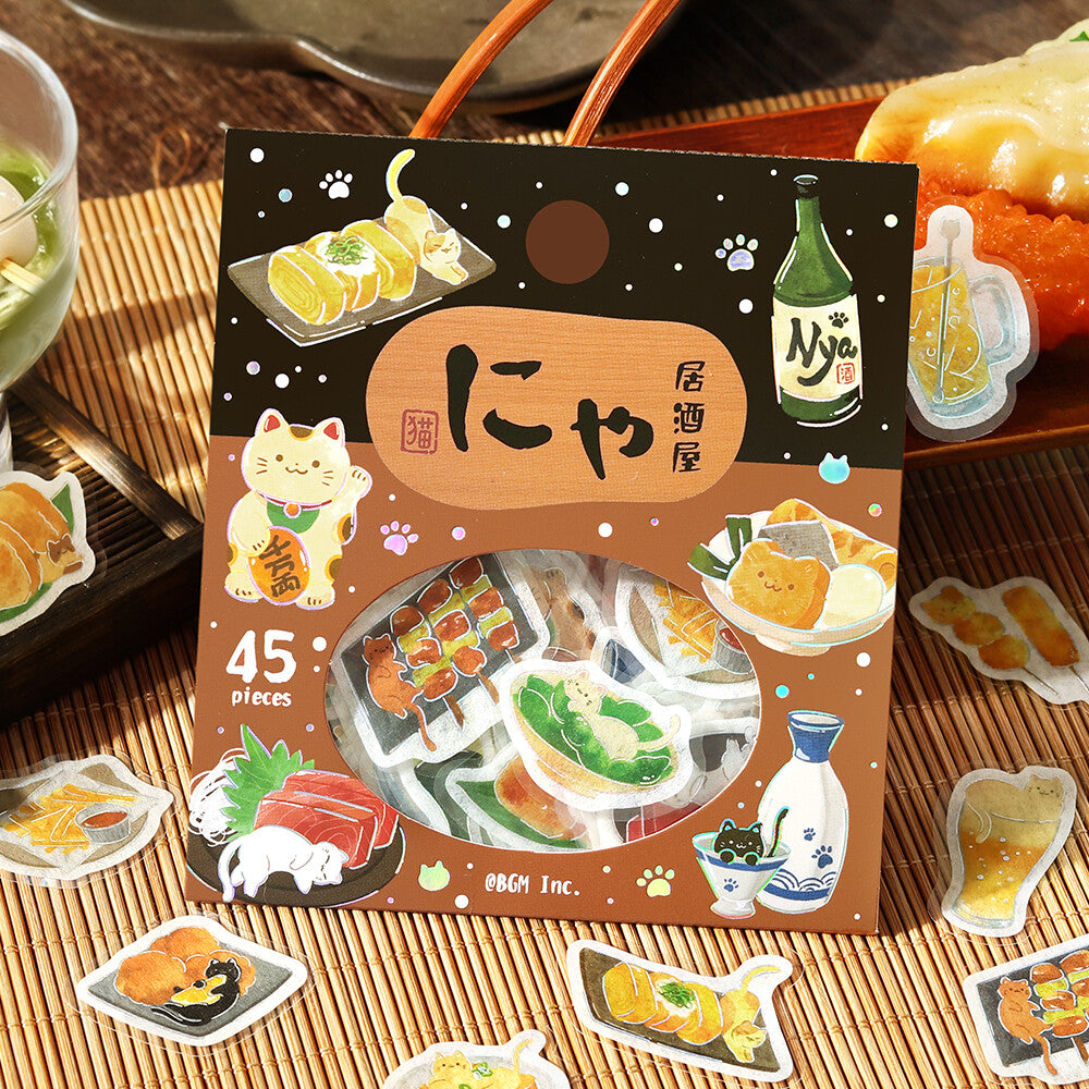 A package of 45 cute cat-themed stickers displayed on a bamboo mat, surrounded by loose sample stickers. The packaging features a dark brown top half and a warm brown bottom half with illustrations of sushi, a Maneki-neko cat, a green bottle, and a beer mug.