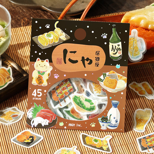 A package of 45 cute cat-themed stickers displayed on a bamboo mat, surrounded by loose sample stickers. The packaging features a dark brown top half and a warm brown bottom half with illustrations of sushi, a Maneki-neko cat, a green bottle, and a beer mug.