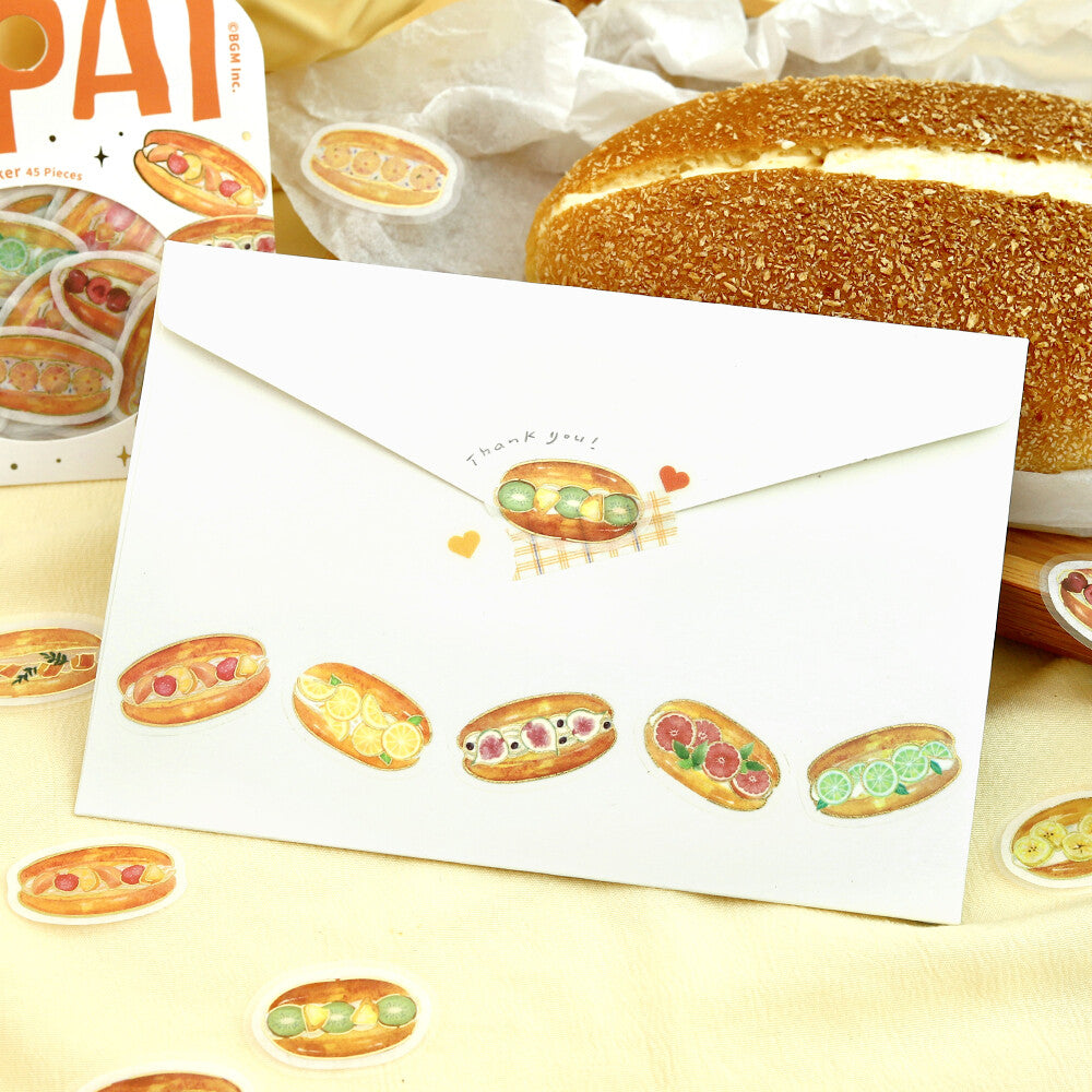 A white envelope decorated with cute watercolor hot dog illustrations and the words 'thank you!' on the flap, styled on a yellow surface with bread and sticker props.