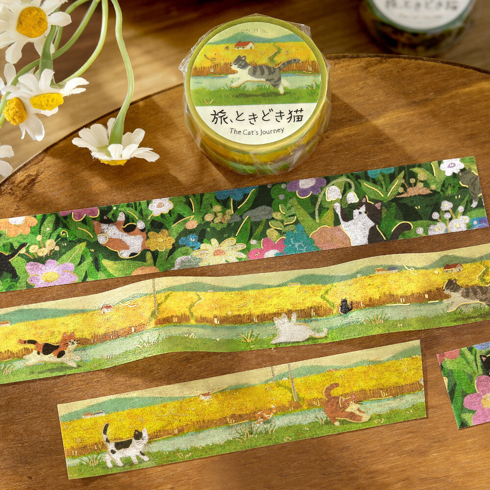 A roll of decorative washi tape labeled 'The Cat's Journey' is displayed on a wooden surface with several strips unrolled. The tape features whimsical illustrations of cats in a yellow field surrounded by green grass and colorful flowers.