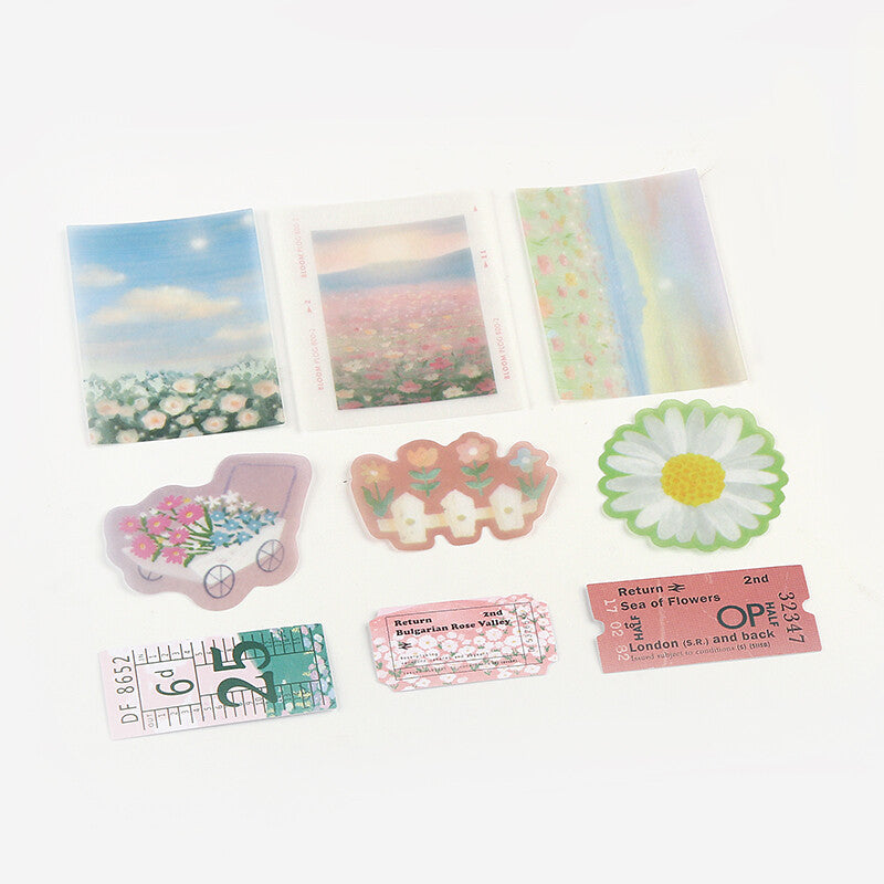 A set of decorative stickers and small printed embellishments arranged on a white background, including floral art pieces, a daisy sticker, a pink flower cart sticker, and ticket-style labels with vintage text.