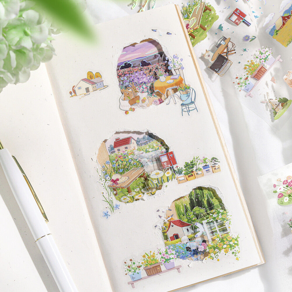 Illustrated stickers of houses and nature on a notebook with a pen and plant in the background.